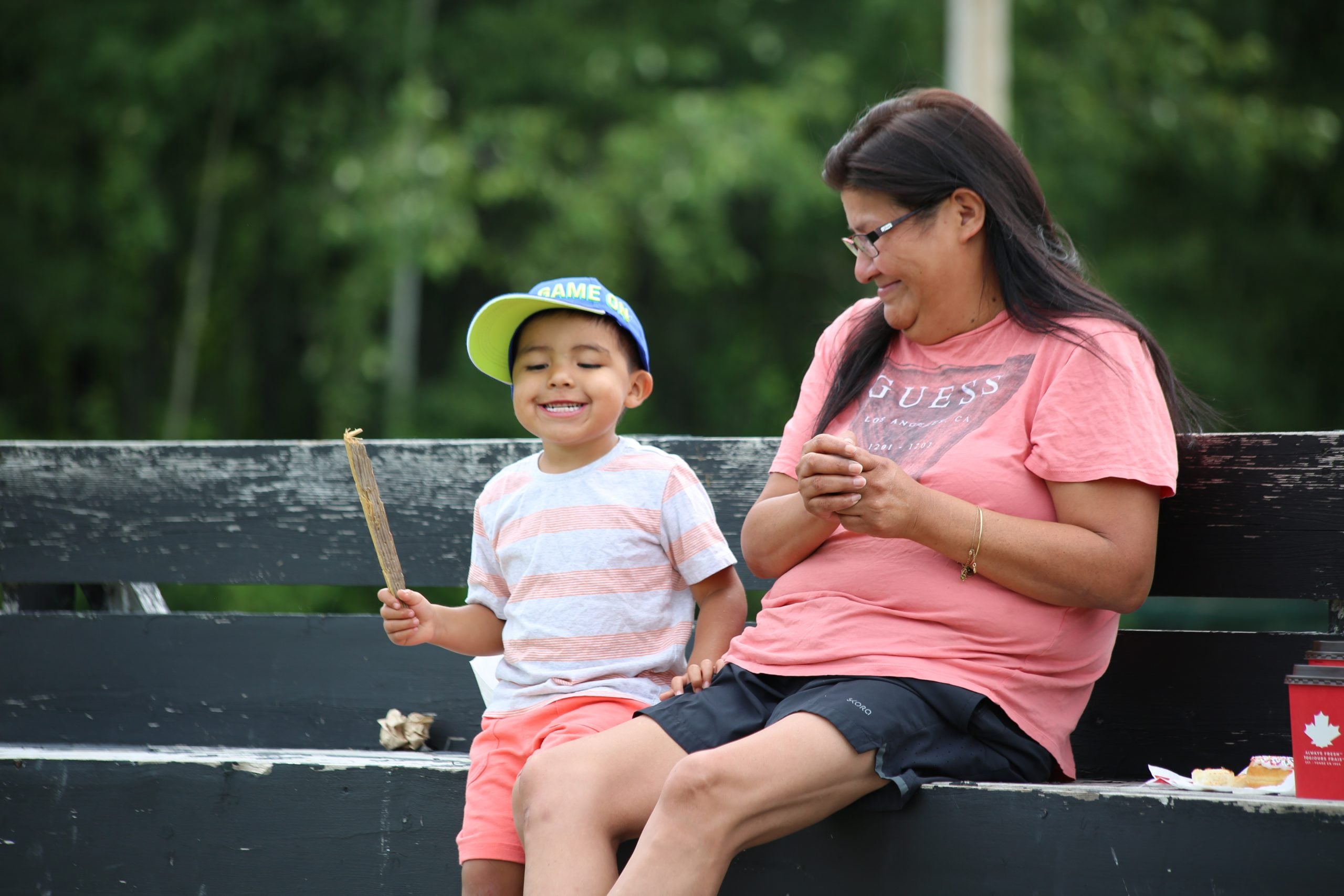 A boy and his mother smiling on a bench. The boy is holding a stick