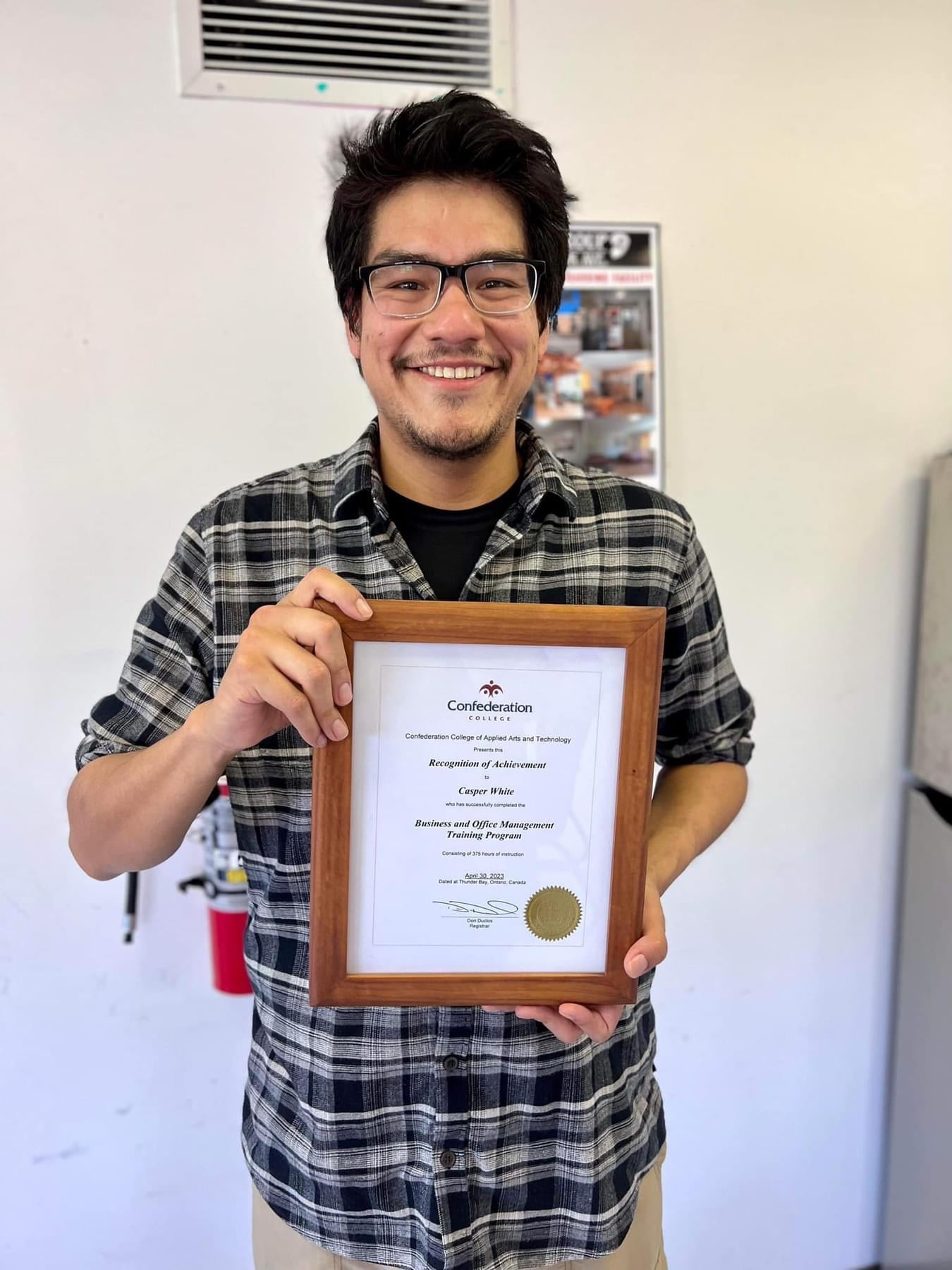 Casper White holding their framed certificate in Business and Office Management from Niiwin Wendaanimok's Training Program. They are wearing a black and white flannel button up on top of a black shirt, and tan trousers. They have glasses and are smiling proudly.