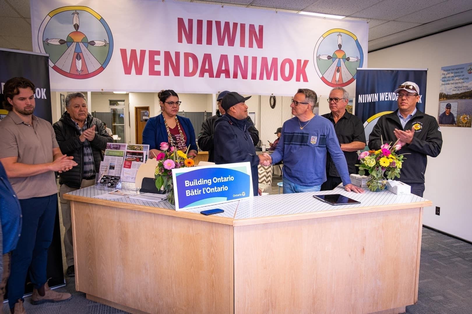 Community members are standing behind a reception desk, with a Niiwin Wendaanimok banner pinned at the ceiling above it. Two people in the middle of the crowd are sharing a celebratory handshake while people applaud.