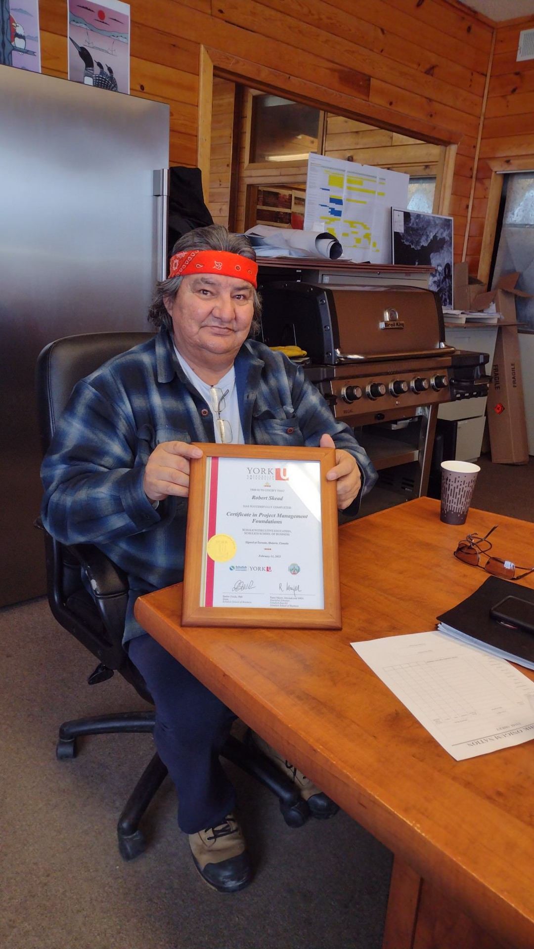 Robert Skead holding their framed certificate in Project Management Foundations from Niiwin Wendaanimok's Training Program. They are wearing a navy flannel with light blue stripes on top of a white shirt, navy pants, and a red bandana tied around their head.